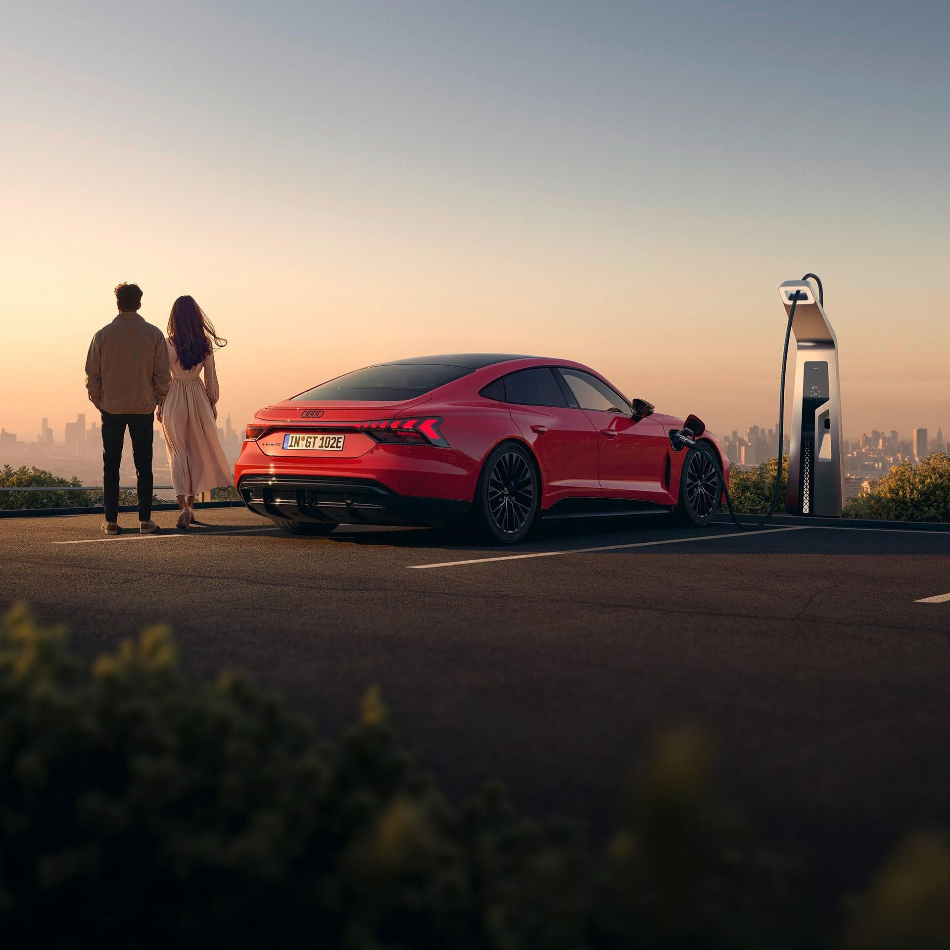Ein Paar steht bei Sonnenuntergang an einem Aussichtspunkt und blickt auf die Stadt, während ihr roter Audi e-tron GT an einer Ladesäule aufgeladen wird.